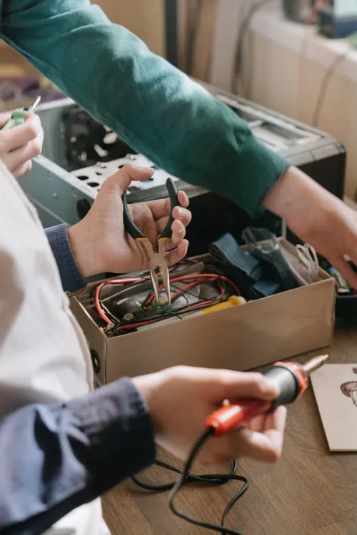 Hände arbeiten mit Zange und Lötkolben an einem geöffneten elektronischen Gerät.