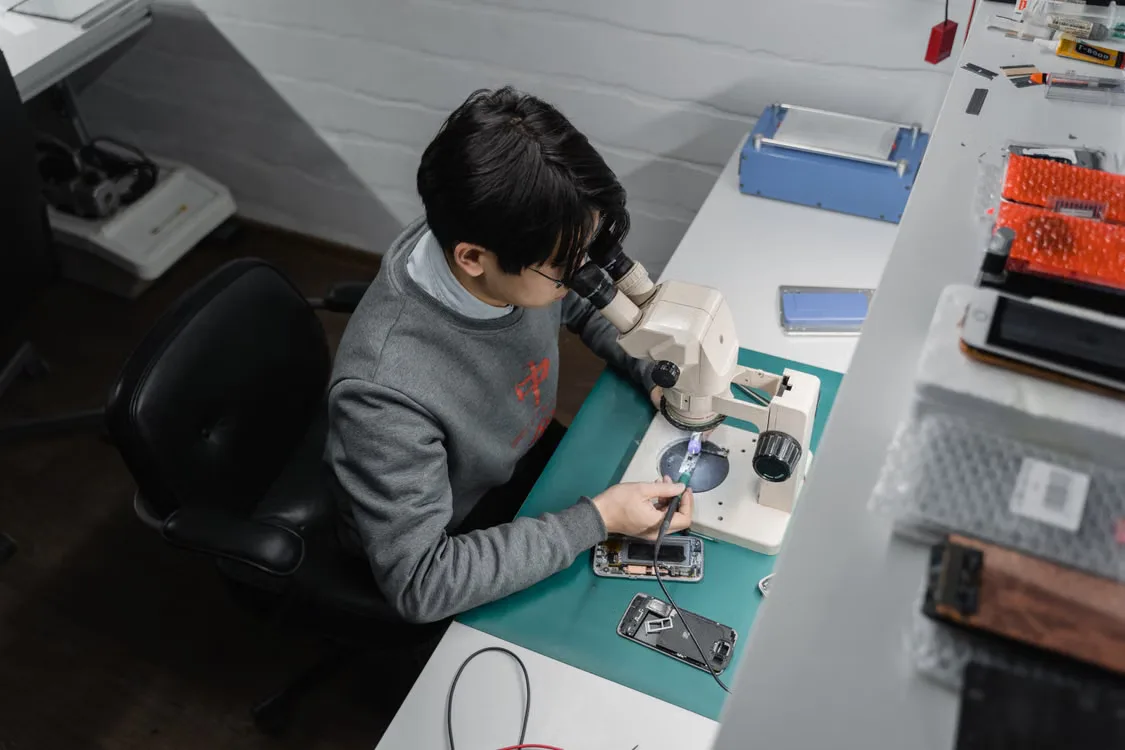 Ein Techniker untersucht ein geöffnetes Smartphone am Werkstatttisch unter einem Mikroskop.